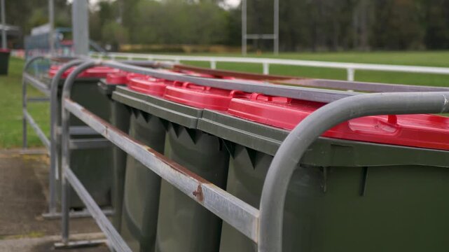 Slow motion close-up of a row of green mobile garbage rubbish bins with red lids lined up behind a metal bar railing next to an outdoor public sports field oval in a rural town suburbs of Australia