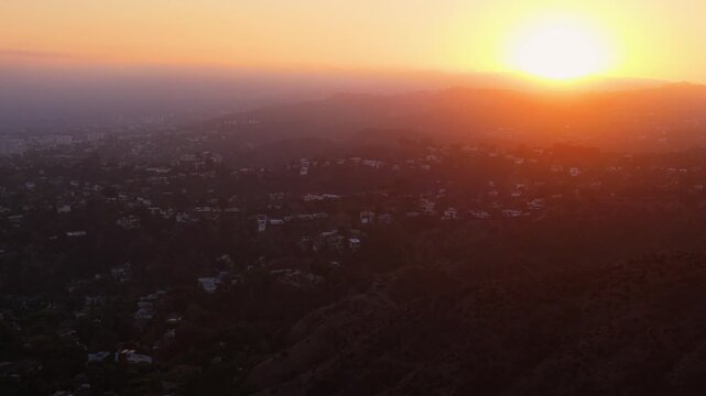 Hillside homes scattered across a dark canyon slope as a blazing sun sinks into thick smog over distant ridges. Aerial drone view of hazy city sprawl at sunset Hollywood, Los Angeles, California