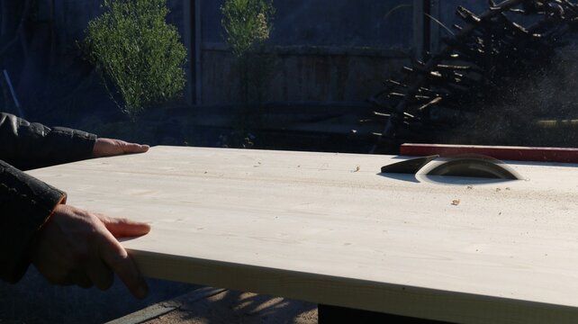 Off-camera, a carpenter carefully guides a wide beech wood panel toward the rotating saw blade of a machine. The process of rip-cutting timber takes place outdoors on a sunny day.