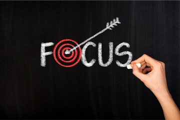 Chalk lettering spelling focus on a blackboard, highlighted by a red target and arrow, with a hand holding chalk, symbolizing goals, concentration, and achievement. © Umm Hamzah