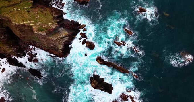 Aerial top-down view of sea waves crashing on rocky cliffs creating white foam texture at Moniello cove, Asturias, Spain.