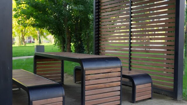 Modern wooden and metal park benches beneath a shaded pavilion in a green urban park. Tbilisi