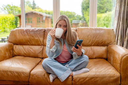 Woman sits on couch, drinks coffee, and looks at phone in a cozy living room setting