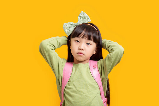 Serious Asian schoolgirl holding head, confused or stressed child with backpack on yellow background, problem and emotion concept
