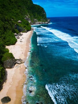 Aerial view of Pantai Nunggalan featuring a pristine sandy beach, turquoise ocean waves crashing against the shore, and lush green cliffs in Kecamatan Kuta Selatan, Bali, Indonesia.