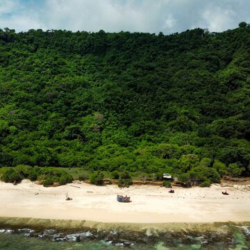 Aerial view of Pantai Nunggalan with a small boat on the white sand beach and dense green forest covering the cliffside in Kecamatan Kuta Selatan, Bali, Indonesia.