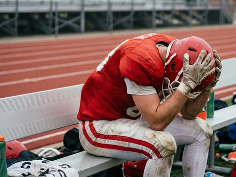Defeated American football player wearing a mud-stained red jersey and white pants sits on a sideline bench with head in hands and helmet on, showing exhaustion and disappointment.