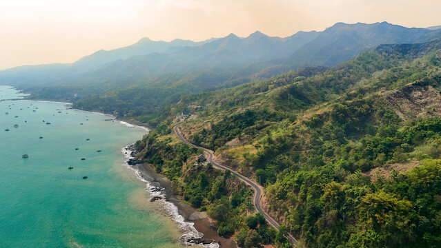 Aerial view of a winding coastal road along the lush green mountainside meeting the turquoise sea in Sukabumi, Indonesia.