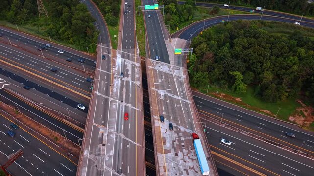 Parallel overpasses with a blue truck above highway lanes. A long vehicle occupies the right bridge deck, while cars pass below and curved exits trace the surrounding tree cover.