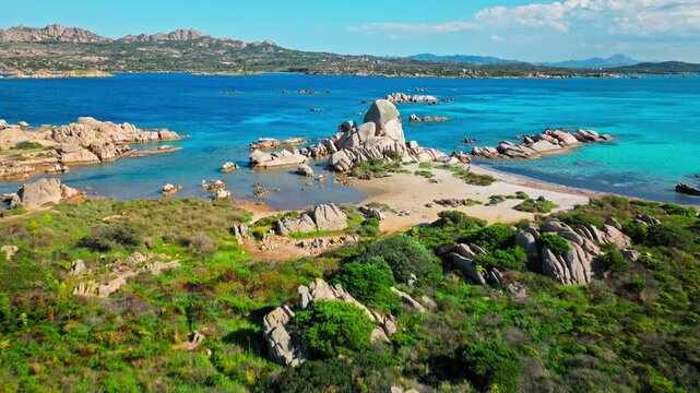 Drone view of Spiaggia Testa del Polpo in La Maddalena. Tourists on the sandy shore, granite rocks and turquoise water create a lively Mediterranean beach scene.