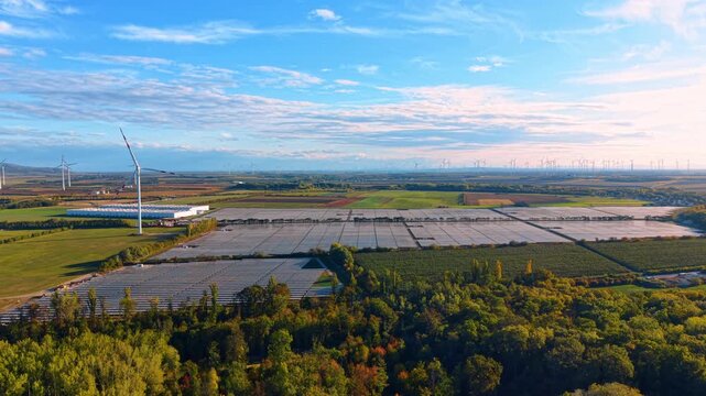 Solar panel arrays and scattered wind turbines across farmland. Broad photovoltaic blocks fill the middle ground behind trees, with crop plots and a bright sky toward the right.