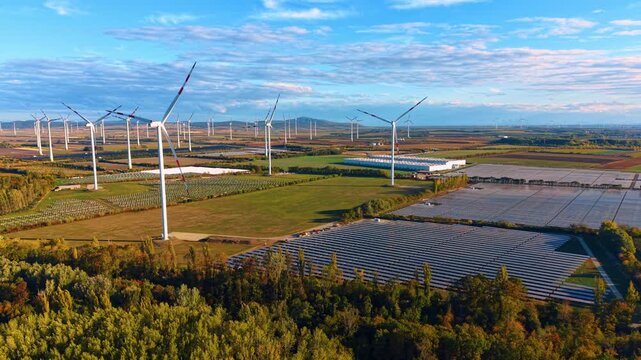 Wind turbines and solar panel rows bordered by woodland. A white rotor rises left of center beside photovoltaic arrays, with green plots, tree belts and distant turbine towers.