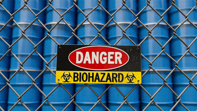 Warnschild Biohazard an Absperrzaun vor gestapelten F&auml;ssern