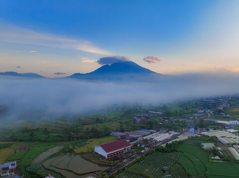 Aerial view of Semayu landscape with rice terraces, greenhouses, and a road with traffic under a mountain peak partially covered by morning fog in Selomerto, Central Java, Indonesia.