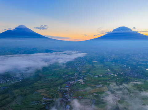 Aerial view of the Semayu area with twin volcanoes rising above lush green terraced rice fields, misty valleys, and soft sunrise light, Selomerto, Central Java, Indonesia.