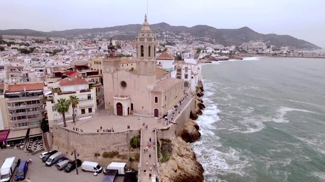 4K drone aerial view of Sitges, Spain, featuring the iconic Sant Bartomeu i Santa Tecla church overlooking the Mediterranean coast, beaches and historic seaside town.