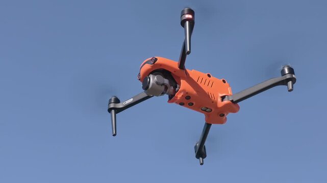 Orange drone hovers in the clear blue sky, displaying its design and aerial capabilities while maintaining a steady position, with propellers visibly spinning and sunlight reflecting off its surface