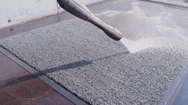 Worker uses a spray nozzle to apply water onto a textured surface, progressively soaking it. The scene captures the transformation as water interacts with the material, enhancing visibility