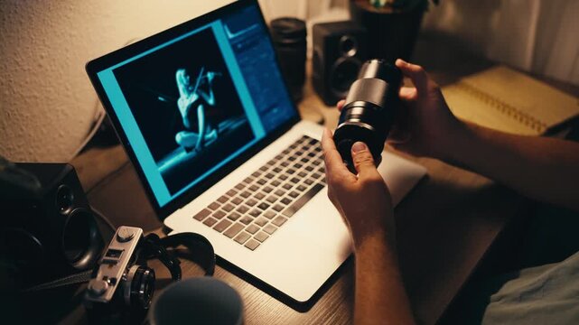 Creative professional photographer examining a camera lens at a warmly lit desk, with a laptop displaying a photo editing software interface and a vintage film camera nearby