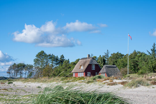 Traditionelles rotes Reetdachhaus hinter wei&szlig;en Sandd&uuml;nen an einer d&auml;nischen K&uuml;ste, umgeben von Strandhafer und gr&uuml;nen B&auml;umen, unter blauem Himmel mit wei&szlig;en Wolken und d&auml;nischer Flagge.