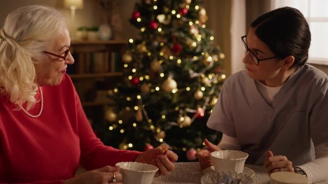Senior Caucasian woman drinking tea with female caregiver near Christmas tree. Footage suits eldercare promotion, geriatric healthcare advertising, holiday companion services