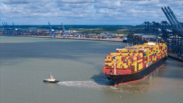 Aerial view of a large container ship being guided by a tugboat into the Port of Felixstowe, with shipping containers and cranes along the docks in Felixstowe, England, United Kingdom.