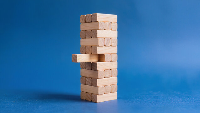 Wooden block tower game with single piece pulled out sideways against blue background, balanced stack of timber bricks, conceptual risk management fragile stability and strategic decision concept