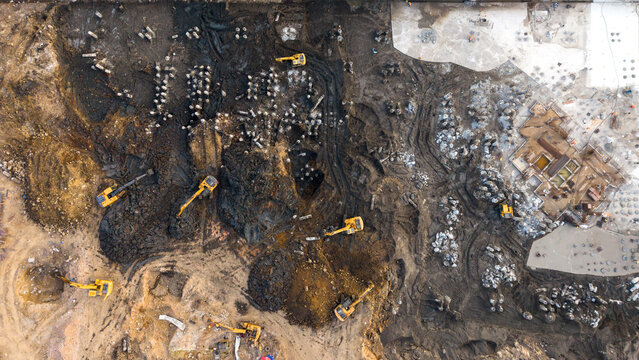 Aerial view of multiple yellow excavators and cranes working on a large muddy construction site with piles of earth and concrete foundations in India.