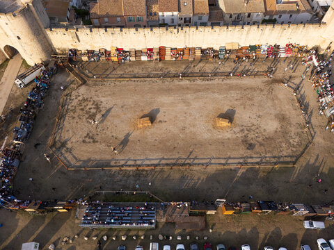 Aerial view of a temporary bullfighting ring set up within the historic stone fortifications, featuring spectators in the stands and golden hour light in Aigues-Mortes, Occitanie, France.