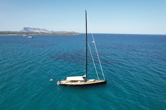 Aerial view of a sleek sailboat anchored in the turquoise waters of the Mediterranean Sea with a mountain coastline in the background in Masua, Sardinia, Italy.