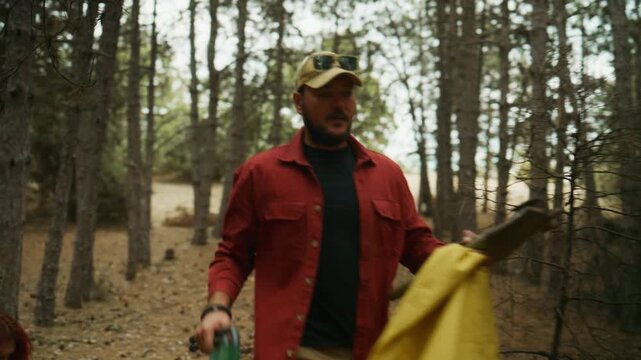 Man collects litter with a trash picker and bag while walking through a pine forest on a dirt path.