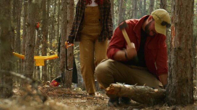 Man chops firewood with an axe while a woman brings a saw to continue cutting wood at a pine forest campsite.