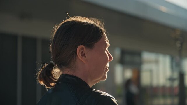 Rear view of a woman sitting on a bench facing an urban plaza as warm late afternoon sunlight shines on her face, 4K