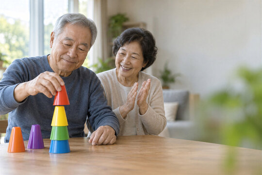 Senior asian man stacking colorful plastic cones while smiling elderly wife claps hands at home table, cognitive therapy activity