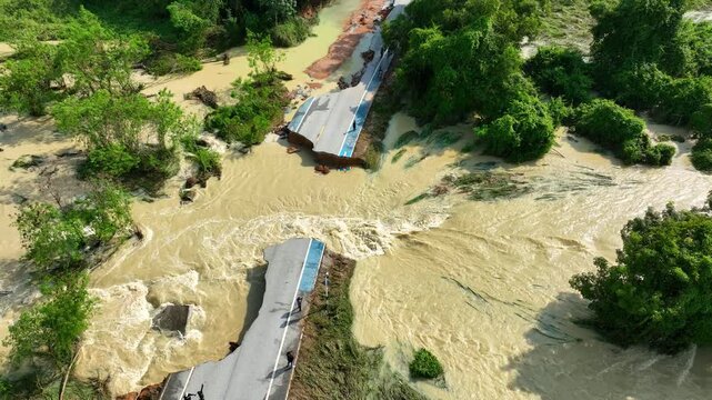 Aerial view of a collapsed rural road with rushing muddy floodwater separating two sections, highlighting infrastructure failure, dangerous travel conditions, emergency response challenges. Thailand.
