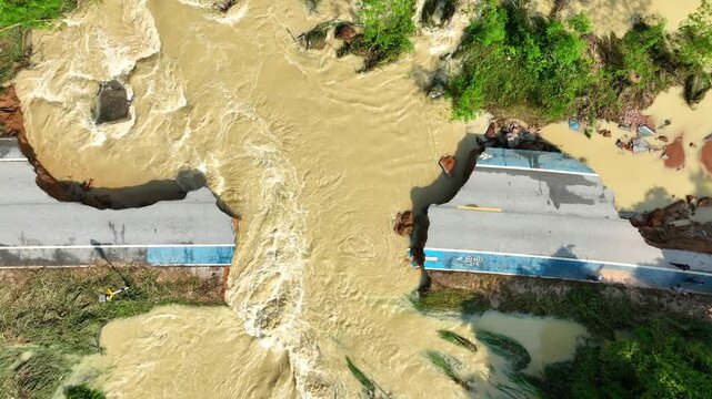 Aerial perspective shows a broken asphalt road washed away by intense flooding, exposing eroded soil and scattered debris while emphasizing the destructive power of extreme weather events. Thailand.
