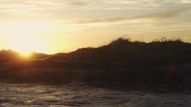 Sun rises beyond a dark cresting shore wave, casting golden light across the ocean surface