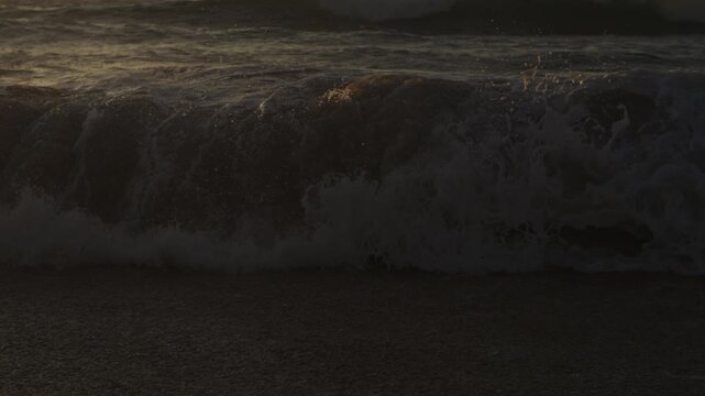 A heavy dark wave folds into white foam near the shore, catching faint warm light along its crest