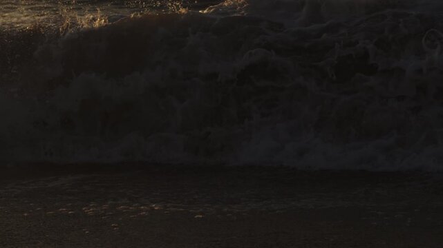 Dense shorebreak foam fills the frame as a dark wave churns beneath warm sunrise light