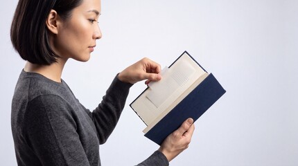 Naklejka premium Focused Young Asian Woman in Gray Sweater Reading and Organizing Pages of a Blue Hardcover Book with a Translucent Bookmark against a Clean White Background for Education and Lifestyle Concept