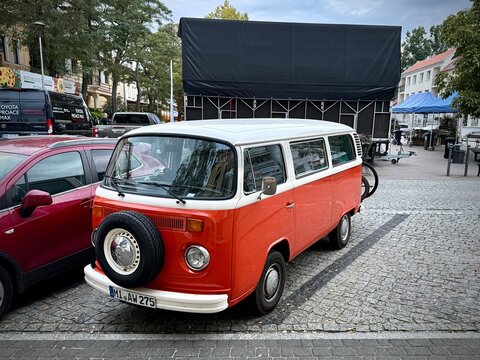 A horizontal photograph capturing a classic orange and white Volkswagen Type 2 (T2) camper van parked on a cobblestone street, showing side profile, front-mounted spare tire, side windows 