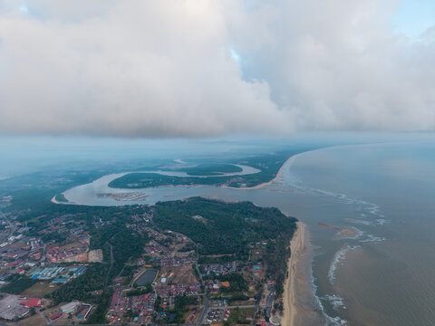 Clouds loom over winding river meeting sea. Coastal town sprawls beside sandy shore and green hills at Rompin
