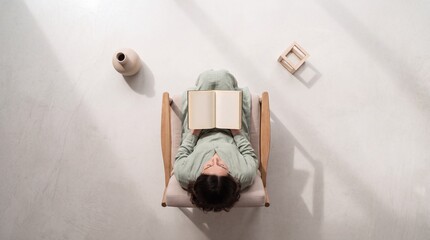 Naklejka premium Top down overhead view of a young woman sitting in a minimalist wooden armchair reading an open book in a bright, clean indoor space with soft natural lighting and long artistic shadows