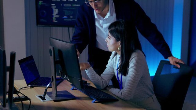 Male and female IT professionals discussing a computer screen in a dark office with blue lighting, collaborating on technical problems, software troubleshooting and digital solutions.