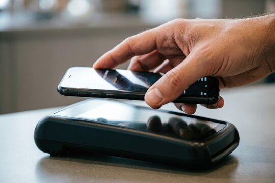 Primer plano de la mano de una persona realizando un pago sin contacto con un tel&eacute;fono inteligente sobre un lector de tarjetas