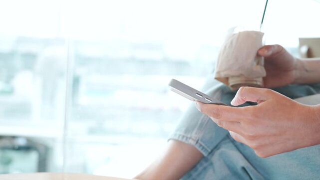 Woman using smartphone while holding iced coffee at cafe, scrolling and texting before typing with both hands, digital chatting, online reading and research during leisure time while waiting.