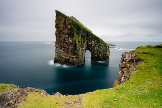 View of Drangarnir sea arch with mossy cliffs and calm ocean waters, Faroe Islands.