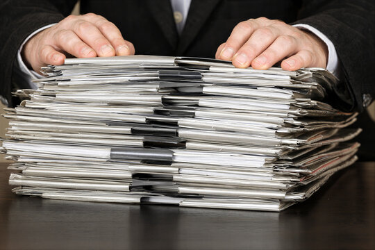 Businessman hands resting on huge stack of folders and paperwork on dark wooden desk close up