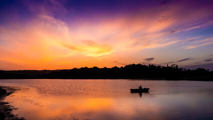 Silhouette of a lone angler fishing from a small boat on a calm lake at sunset, with a vivid orange, pink and purple sky reflected in the water, Colne, Lancashire, England © Jon