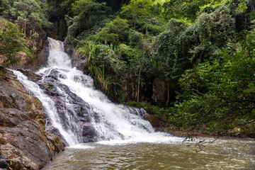 Lush tropical waterfall cascades over mossy rocks into a forest pool framed by dense green foliage and trees. A serene natural landscape evoking tranquility, outdoor adventure. Thac Datanla, Vietnam © evannovostro
